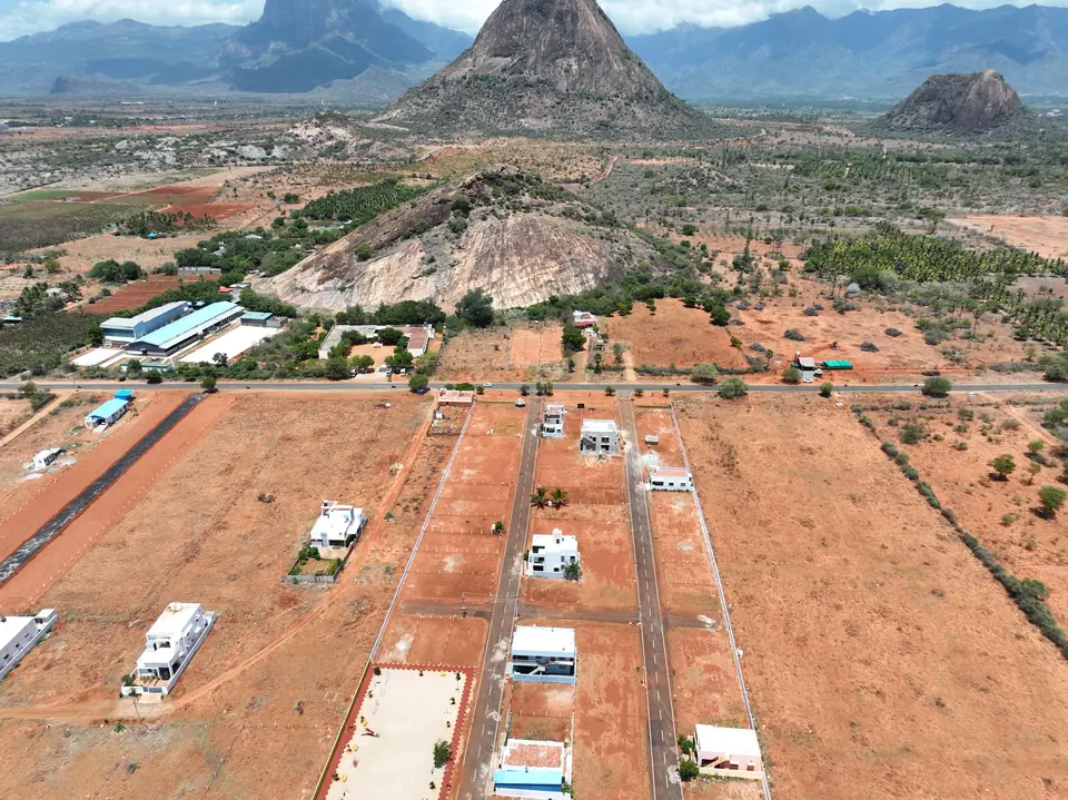 Aerial view of a plotted land development: laid access roads, demarcated plots and a few completed houses, with a conical hill on the horizon